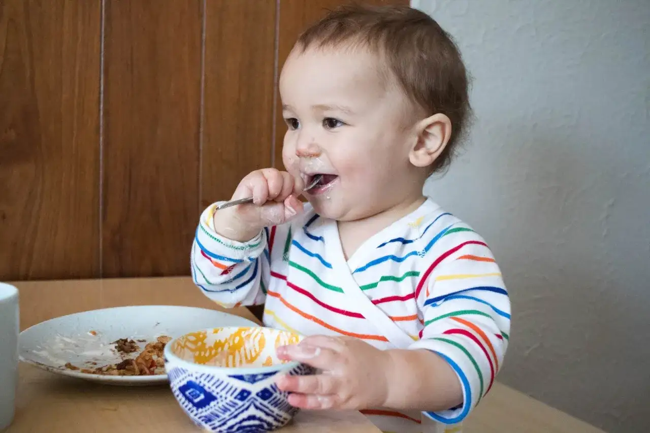 A happy toddler, around 1 year old, is learning at what age a child can eat with a fork, making a delightful mess.