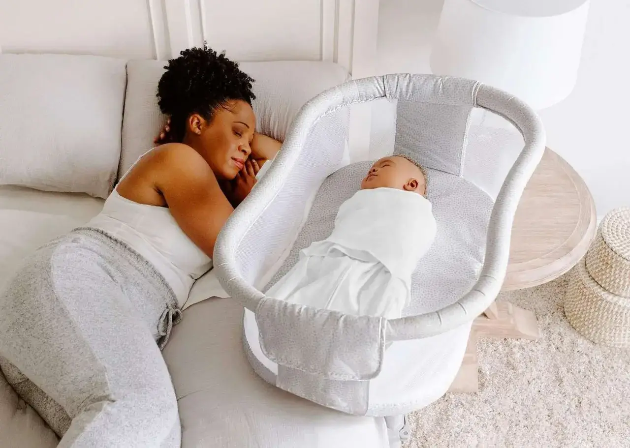 A mother rests beside her sleeping baby in a bassinet, showing what is a bassinet used for: a safe, cozy space for newborns to sleep near parents.