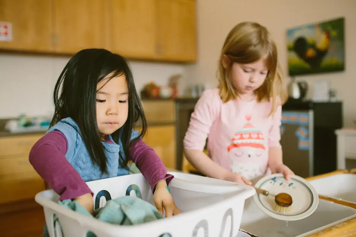 Two young girls engage in child-led learning, washing dishes and clothes in a kitchen setting.