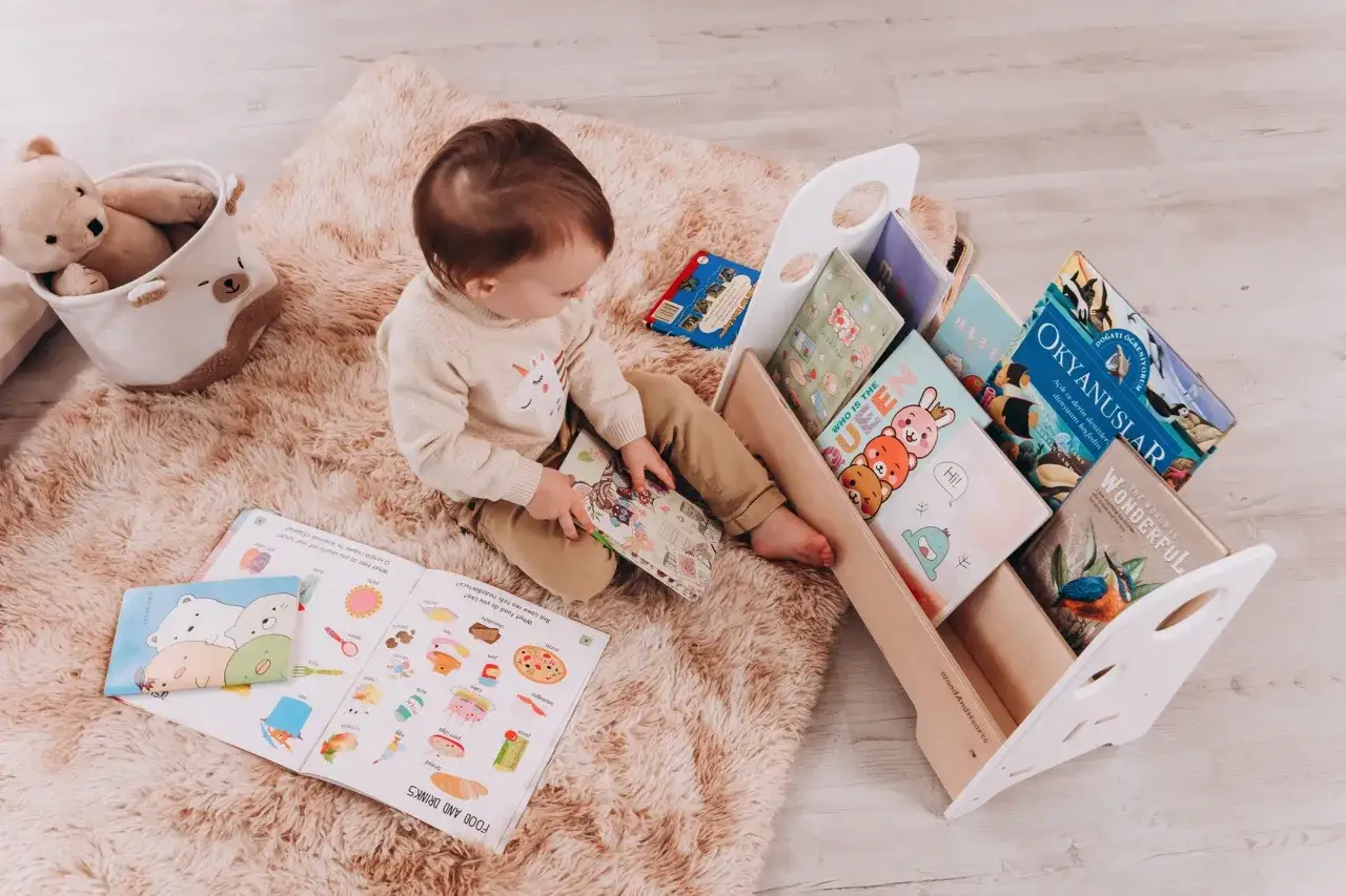 A curious toddler explores a book about food and drinks, surrounded by a collection of colorful books in a wooden Montessori shelf.