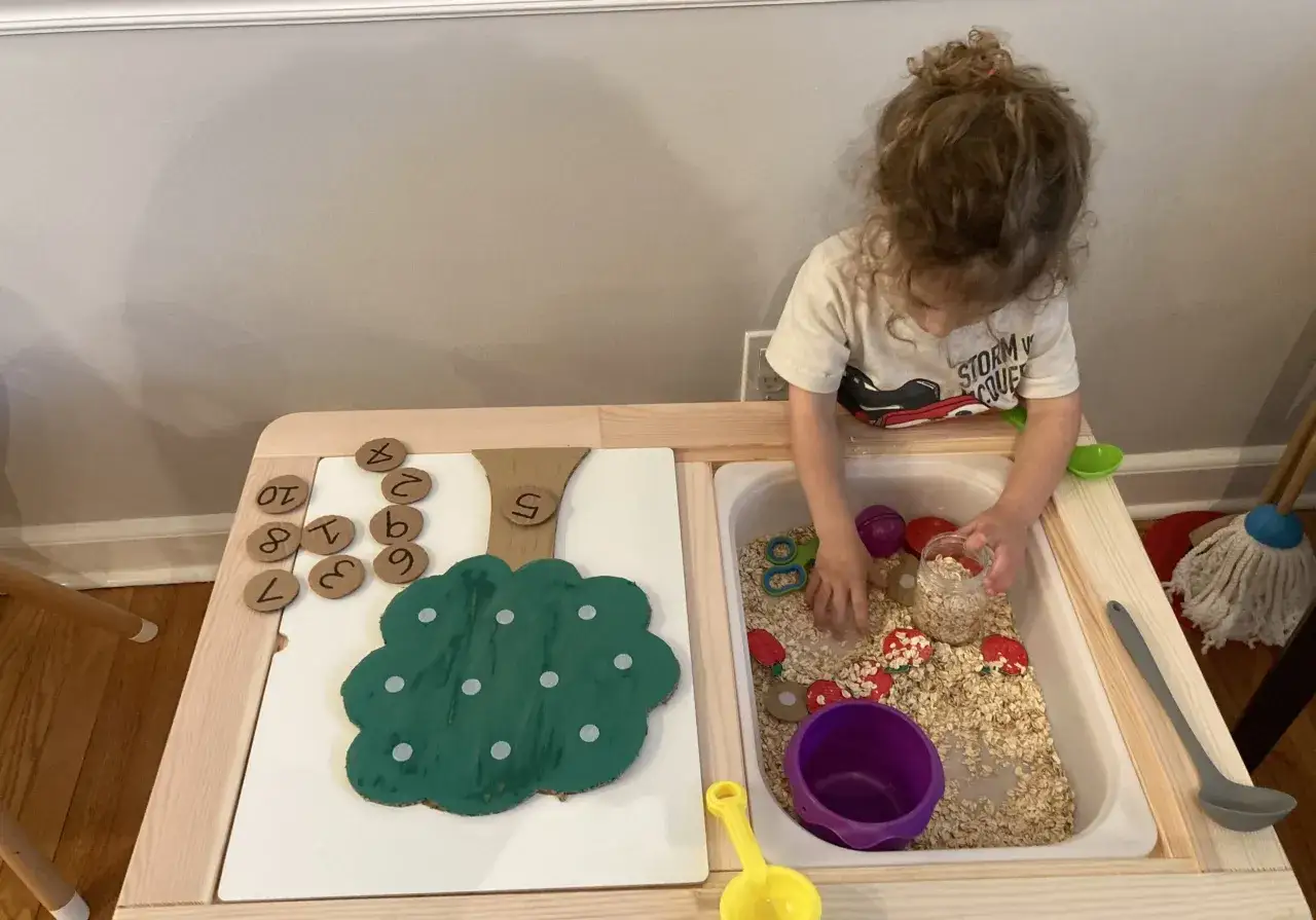 A child plays with a fall sensory bin filled with oats, red apple cutouts, and number tokens.