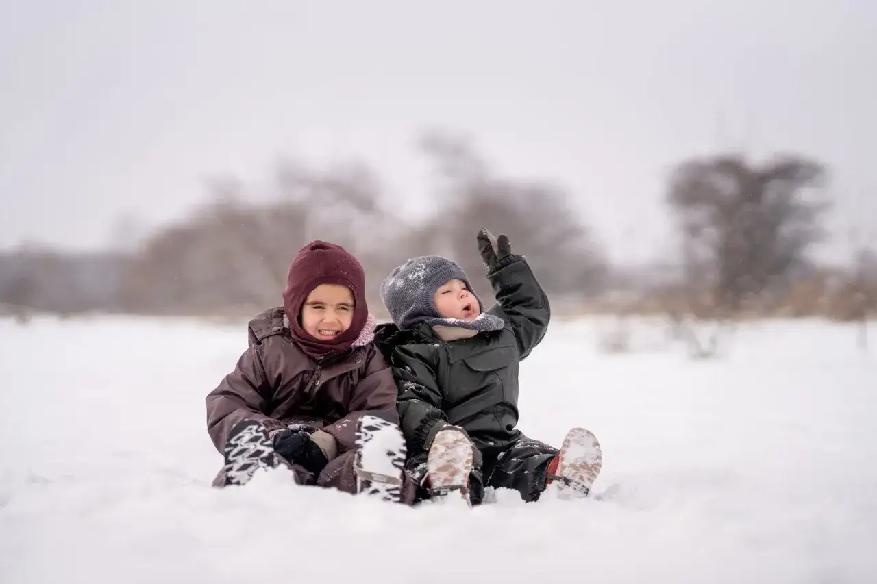 Two children in winter gear sit in the snow, enjoying outdoor fun. Great things to do with a baby in winter include playing in the snow!