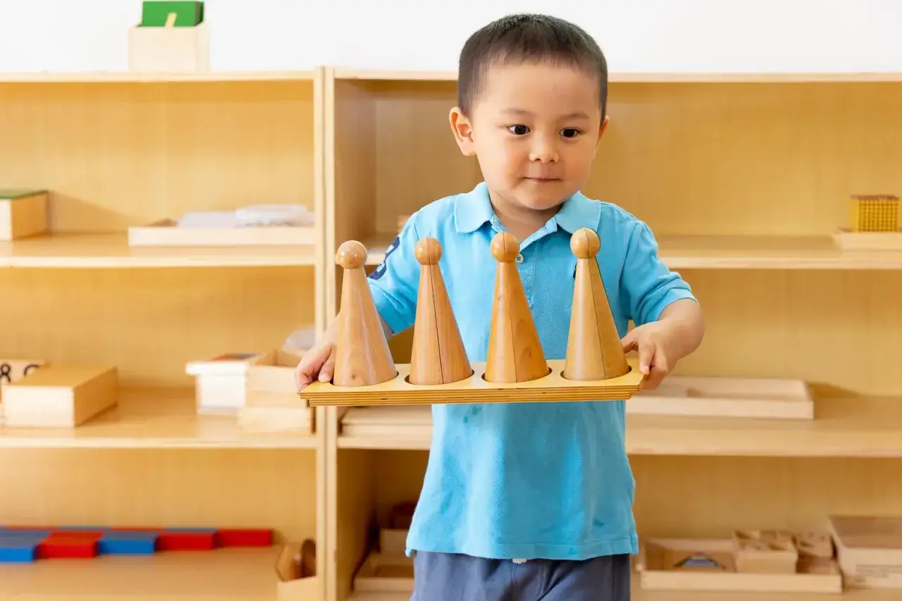 A young boy in a blue shirt holds a wooden tray with four graduated cones, learning about size and order, a key part of Montessori rules.