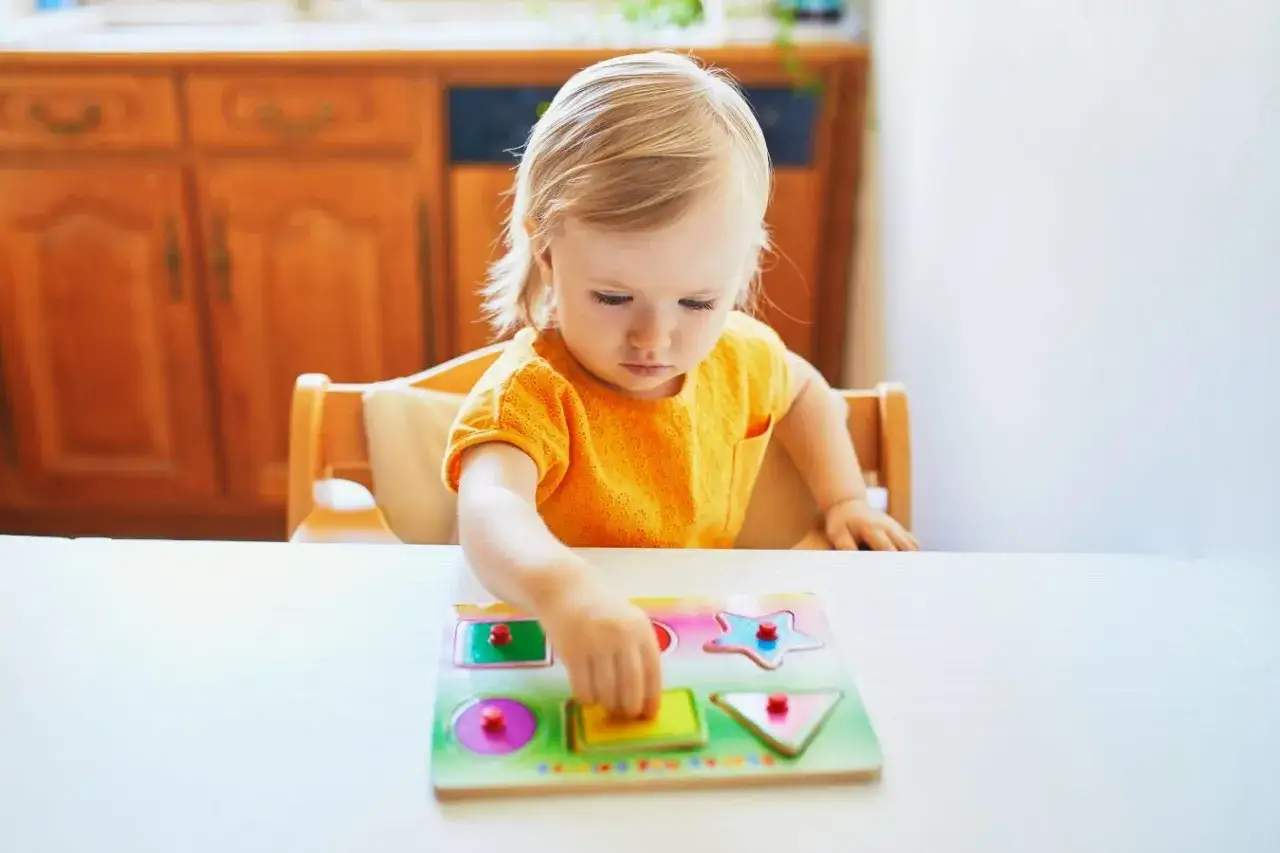 A toddler in an orange shirt concentrates on her first puzzle, a wooden shape sorter with colorful pieces.