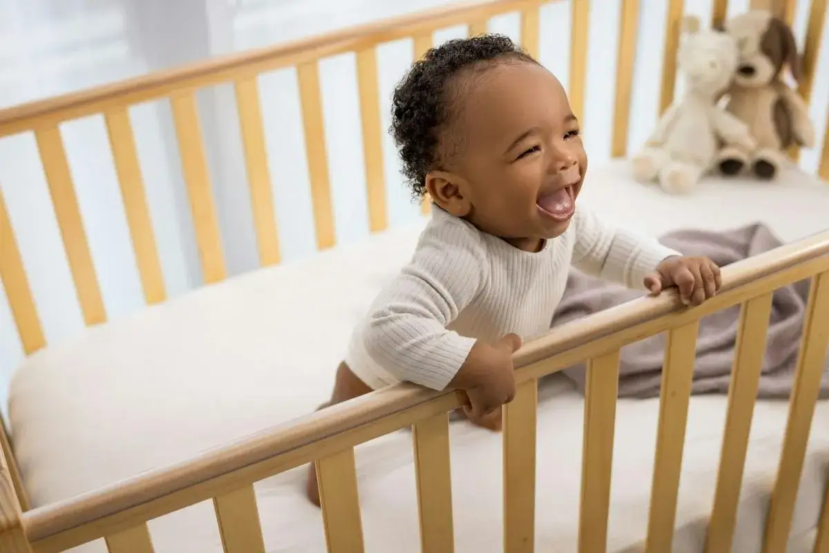 A happy baby laughs while holding onto the rails of a mini crib, showing they are safe and fun.