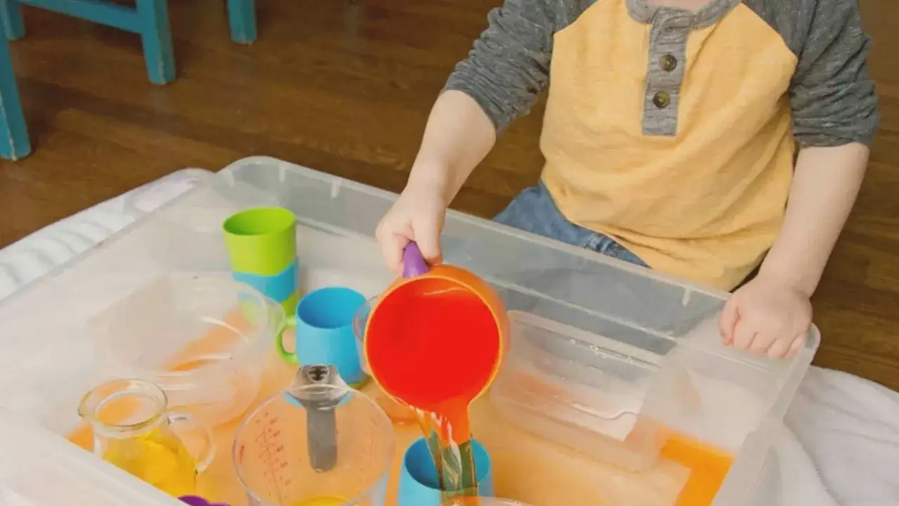 A child enjoys Montessori water play, pouring orange liquid from a scoop into a blue cup.
