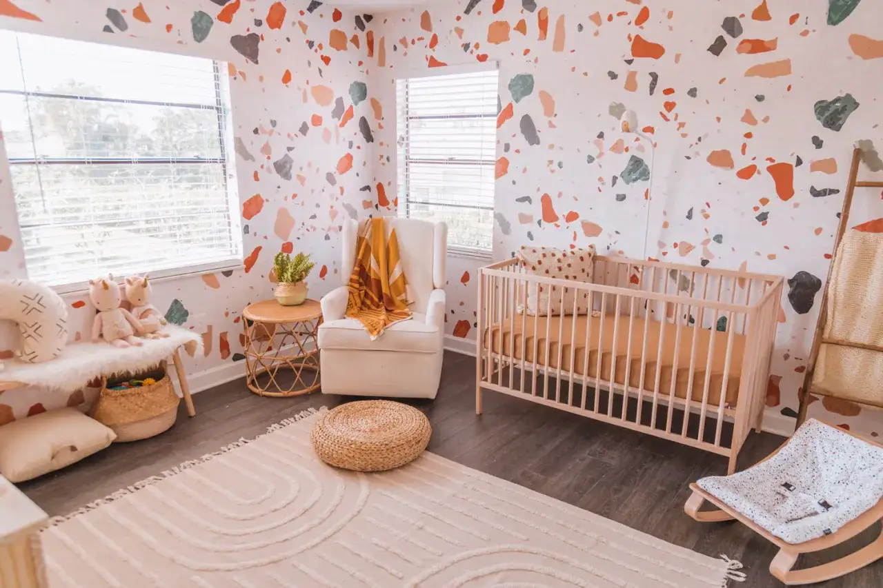 A playful baby girls bedroom with terrazzo wallpaper, a wooden crib, plush toys, and a cozy armchair.