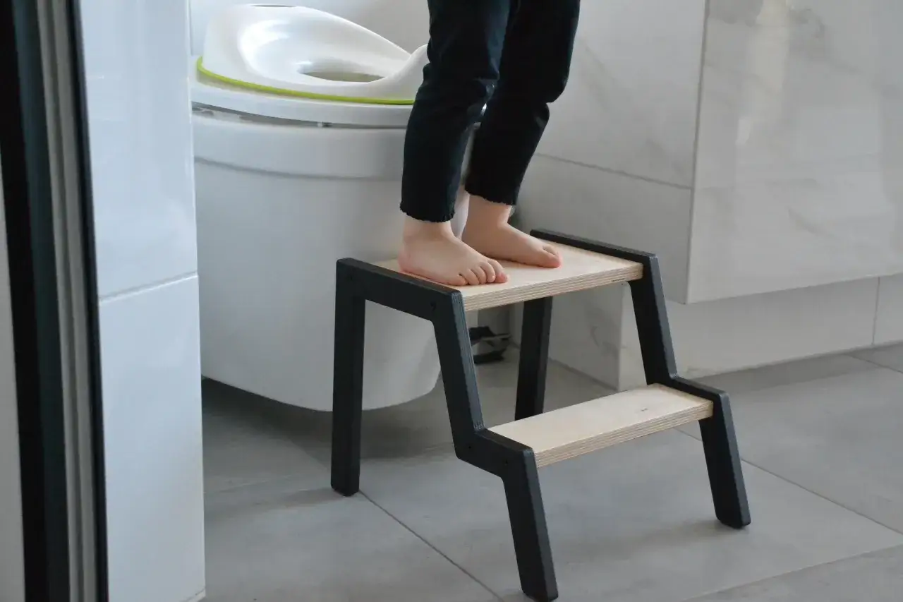 A child's bare feet stand on a two-step stool in a Montessori bathroom, ready to use the toilet.