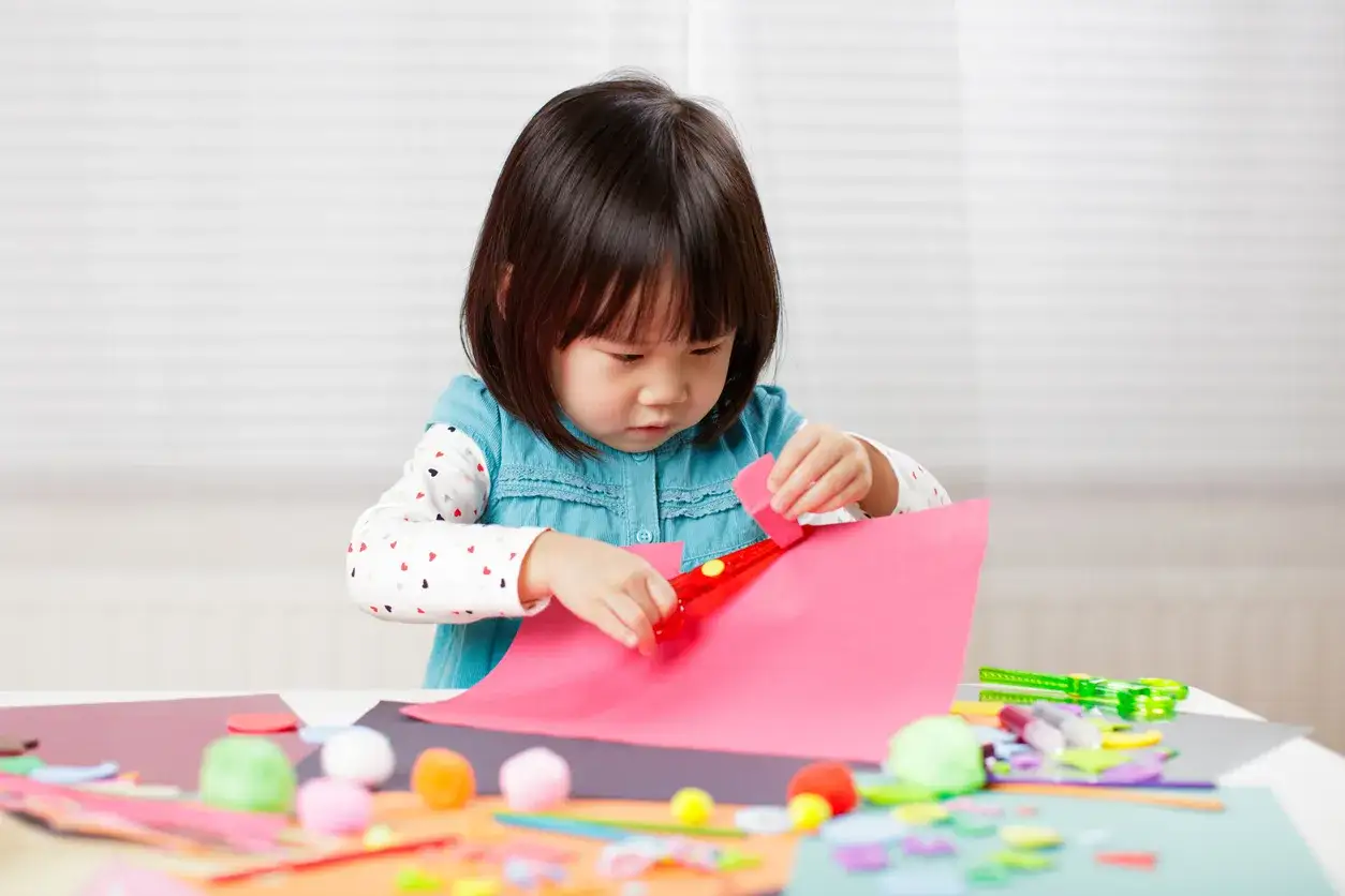 A focused toddler cutting pink paper with red scissors, surrounded by colorful craft supplies.