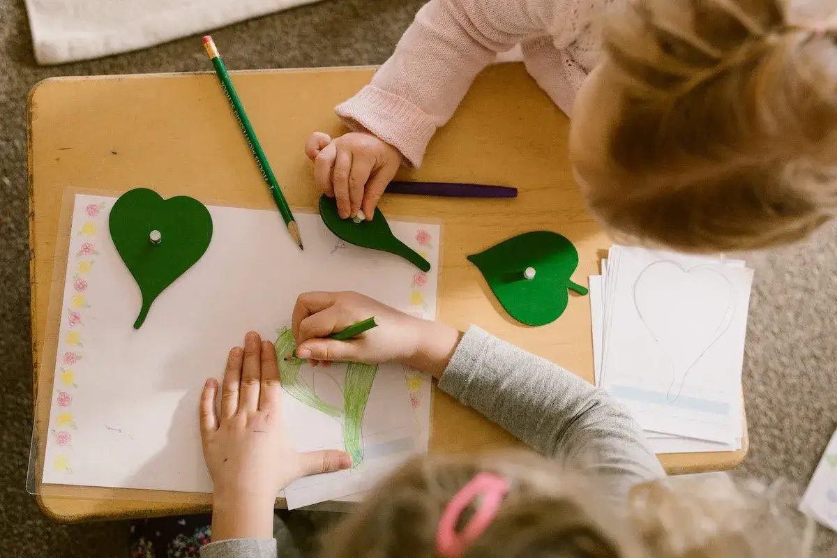Children engage in Montessori fine motor activities, drawing and tracing heart shapes with pencils and green leaf templates on a wooden table.
