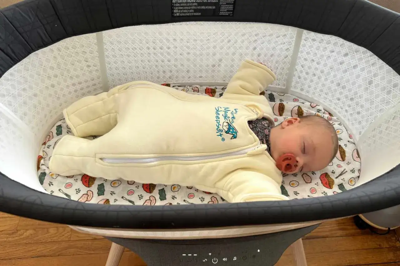 A baby sleeps soundly in a bassinet, proving that babies can sleep in a bassinet. The baby is wearing a yellow sleepsuit with a cute design.