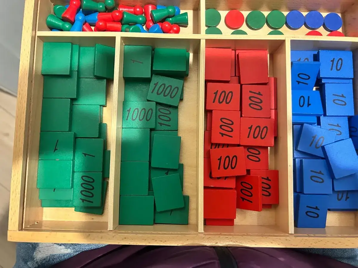A Montessori math set with wooden tiles for play. Green tiles show 1 and 1000, red tiles show 100, and blue tiles show 10.
