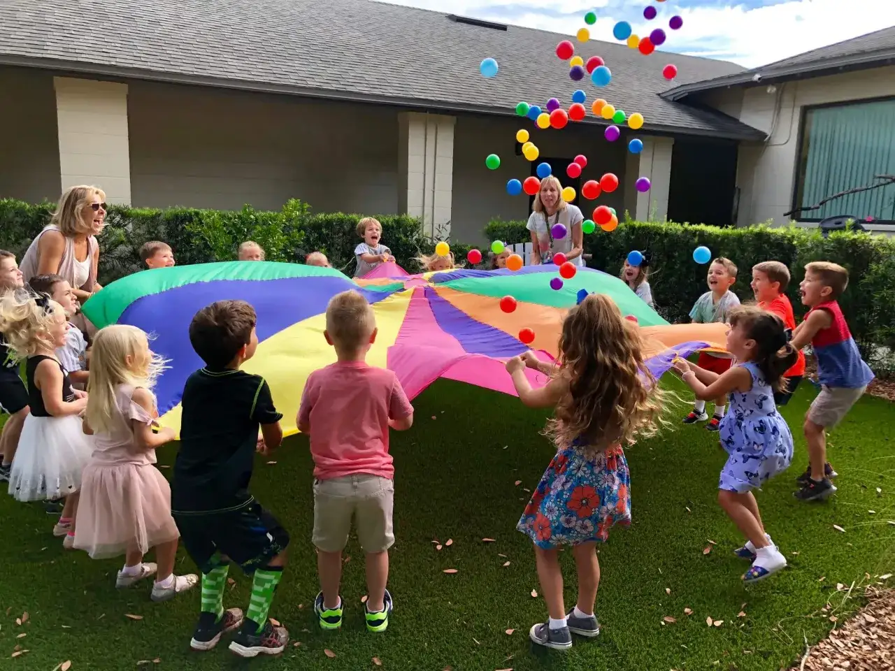 Children joyfully engage in gross motor outdoor activities, tossing colorful balls in the air while holding a large parachute.