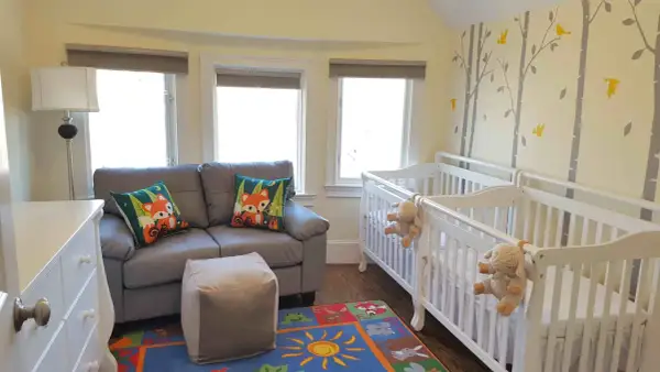 A charming nursery with two white cribs, perfect for nursery sets for twins. A grey sofa with fox-themed pillows and a colorful rug complete the cozy space.
