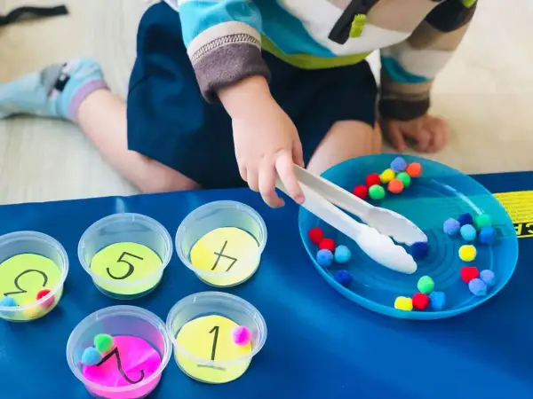 Toddler sorting colorful pom-poms into numbered cups. A fun activity for what to do with toddlers at home, practicing fine motor skills and number recognition.