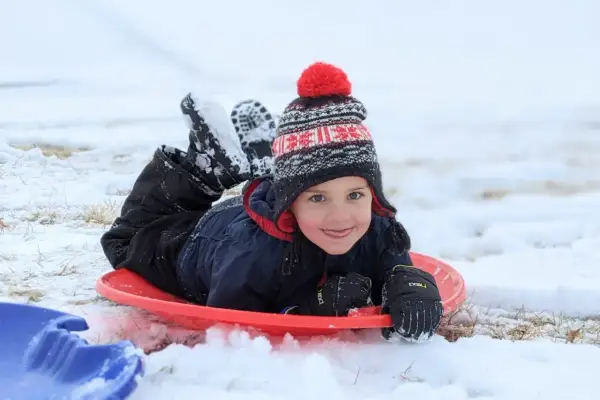 A happy child in a winter hat and snowsuit lies on a red sled, ready for fun. Perfect for things to do with baby in winter, like sledding!