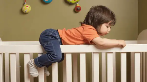 A toddler in jeans and an orange shirt is climbing over the side of a crib, with their legs dangling, as if preparing for dropping crib mattress to floor.