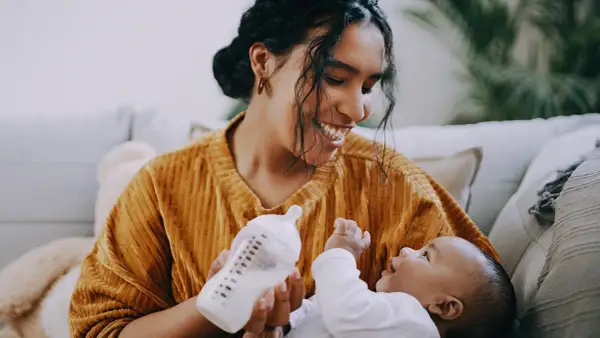 A mother smiles while paced feeding her newborn with a bottle. The baby looks up at her, reaching out a hand.