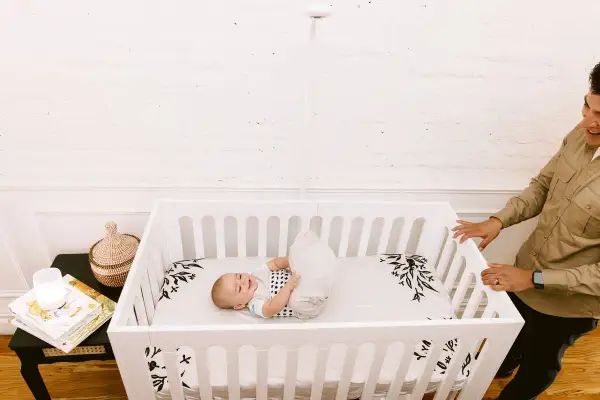 A happy baby rolls around in a white baby crib twin. A man smiles as he looks into the crib.