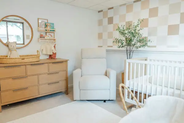 Cozy and modern nursery with a wooden dresser, plush recliner, and crib. This room offers sweet, cute baby room ideas with its neutral palette and playful checkered wallpaper.