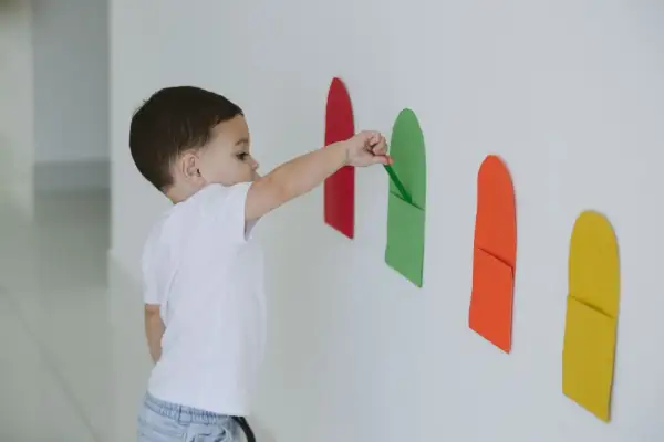 A toddler explores bright colors for 1 year old, placing a green paper strip into a matching pocket on a white wall.