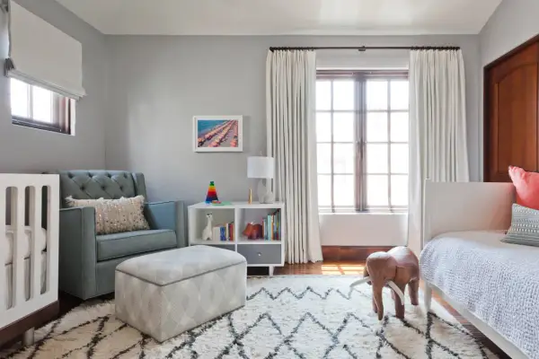 A cozy nursery features a tufted blue couch, a crib, and a white bookshelf filled with toys and books.