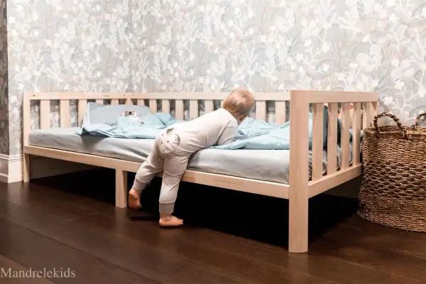 A toddler climbs into a Montessori bed, showcasing its low profile and ideal dimensions for independent exploration.