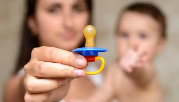 A mother holds a colorful pacifier, perhaps contemplating extended pacifier use for her baby, who is blurred in the background.
