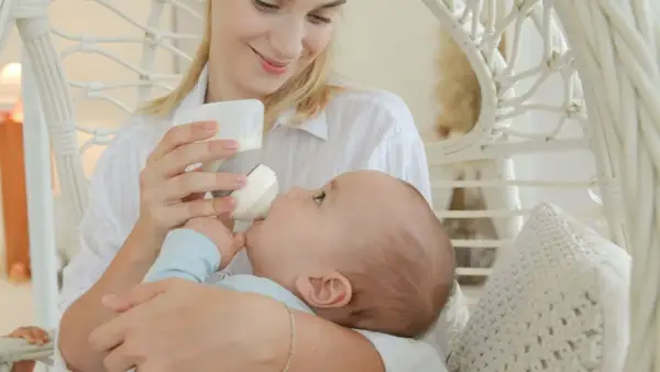 A mother smiles lovingly while holding her baby for upright bottle feeding. The baby is nestled in her arms, taking milk from the bottle.