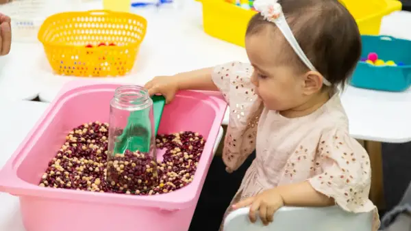A baby girl explores a winter sensory bin filled with beans and a scoop.