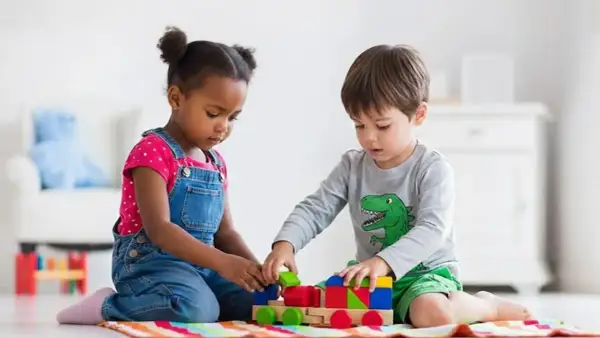 Two children engage in parallel play, building a colorful wooden train with blocks.