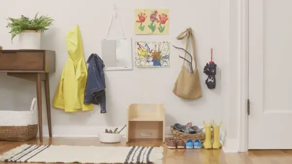 Montessori entryway ideas: a yellow raincoat, denim jacket, and artwork hang on the wall. A small wooden stool and baskets hold shoes and boots.