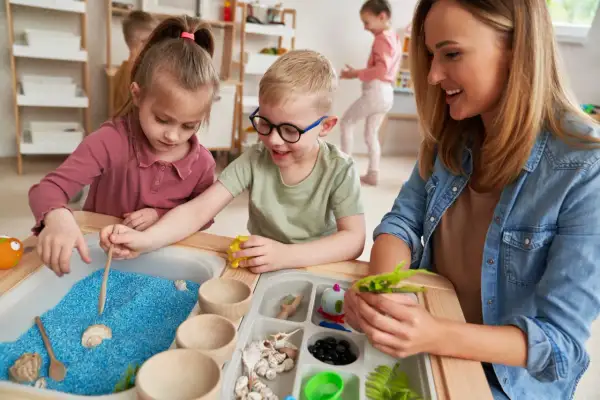 Children explore blue sand and shells in a sensory table, guided by a smiling teacher.