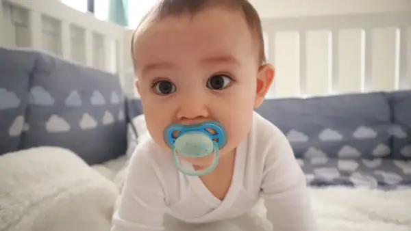A baby with a blue orthodontic pacifier in a crib. Are orthodontic pacifiers better for developing smiles?