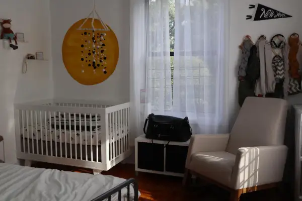 Cozy nursery nook ideas: a white crib, a comfy armchair, and a wall decorated with a sun mural and a New York pennant.