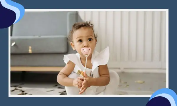 A baby girl with a pink pacifier in her mouth sits on the floor, her eyes wide with curiosity. The pacifier terth is a comforting presence as she explores her surroundings.
