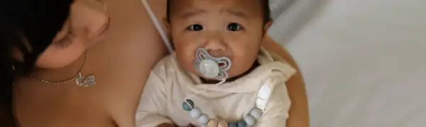 A baby with a pacifier, possibly experiencing pacifier nipple confusion, looks into the camera while being held by a parent.