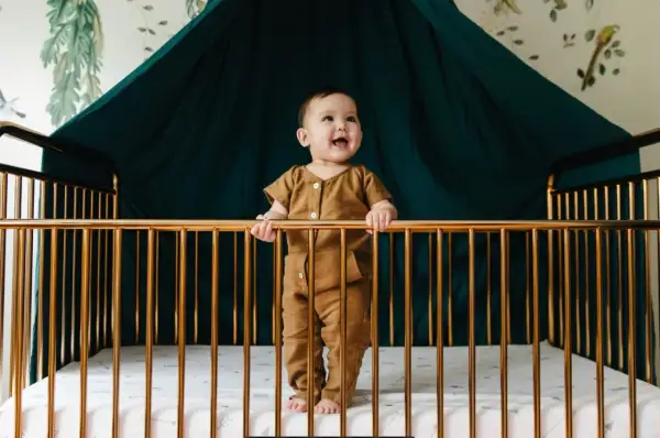 A happy baby stands in a golden crib, holding the bars. What do you need for a crib? A safe space, a soft mattress, and a smiling baby!
