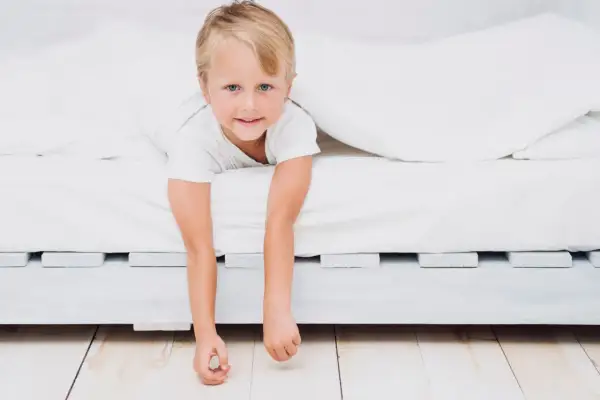 A young boy peeks out from under white sheets, his arms dangling. He looks ready for a new adventure, perhaps signaling it's time to transition to a floor bed.