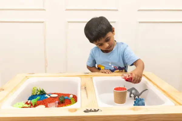 A boy pours red liquid into a sensory table with dinosaurs and colorful rice, exploring fun sensory table ideas.