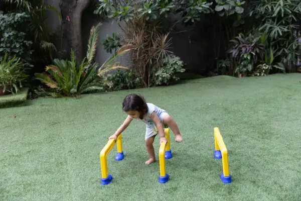 A toddler practices gross motor outdoor activities, balancing on yellow and blue hurdles in a grassy yard.