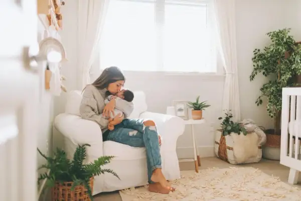 A mother cradles her baby in a cozy nursery, surrounded by soft decor and plants, hinting at peaceful nursery themes.