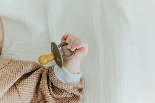 A tiny hand holds a pacifier that appears too big for a newborn, nestled in a soft, textured blanket.