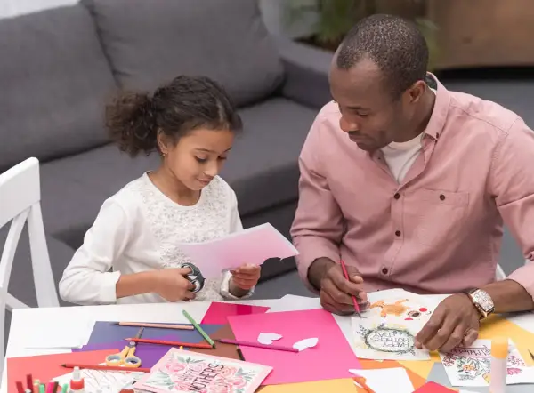 A father and daughter are crafting Mother's Day cards. The toddler cutting paper with scissors is focused on her task.
