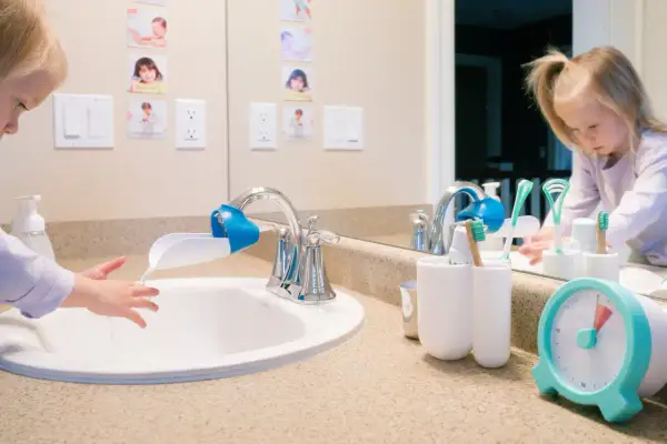A child washes hands at a sink with a faucet extender, in a Montessori bathroom. Toothbrushes and a timer are nearby.