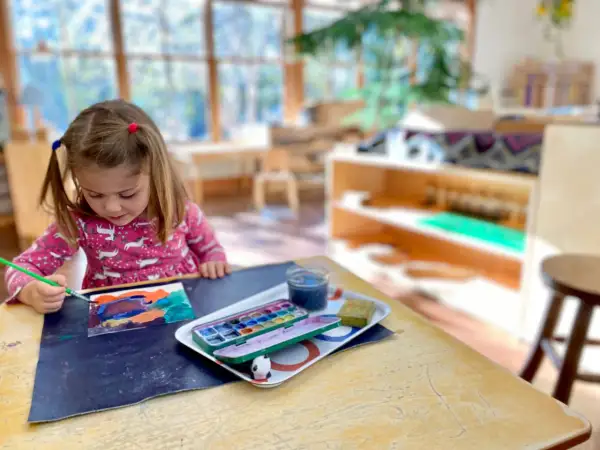 A young girl with pigtails paints at a table, exploring her creativity with watercolors. This scene captures the essence of Montessori nature-inspired learning.