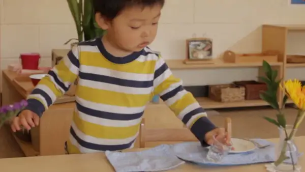 A young child, around 18 months old, practices setting a table in a Montessori environment, carefully placing a glass.