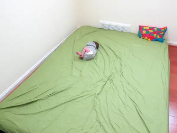 A baby sleeps on a green mattress on the floor, a simple setup for how to make a floor bed for baby. A colorful map pillow rests nearby.