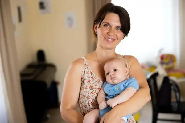 A smiling mother holds her 2-month-old baby, celebrating precious milestones. The baby wears a blue onesie with a yellow and blue heart.