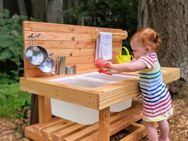 A toddler engages in Montessori movement activities, exploring a wooden mud kitchen with pots and a watering can.